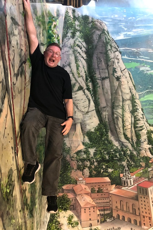 An image of a man who looks like he is hanging from a cliff over Montserrat Monastery, a typical picture of the sort you take in the Museum of Illusions in Barcelona. Photo © Dugald Steer 2024.